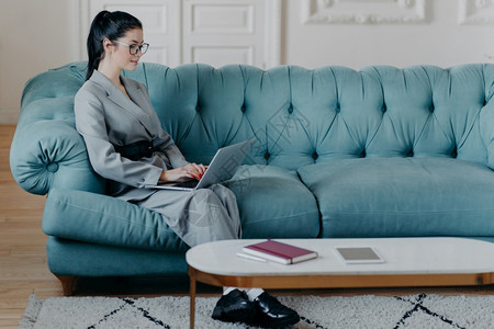 在笔记本电脑上忙碌的女自由职业者键盘坐在舒适的软沙发上正式穿好衣服进行项目通过在线视频聊天与雇员沟通远程工作概念图片