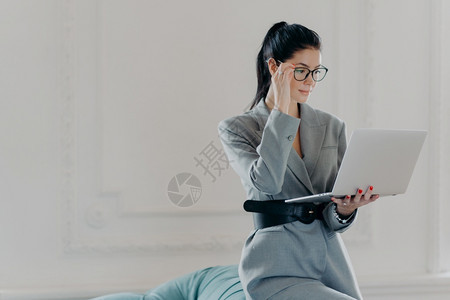 精美的专业女银行家集中在笔记本电脑屏幕上身着正式服装在宽敞的房间家庭办公室阅读在线版本的文件图片