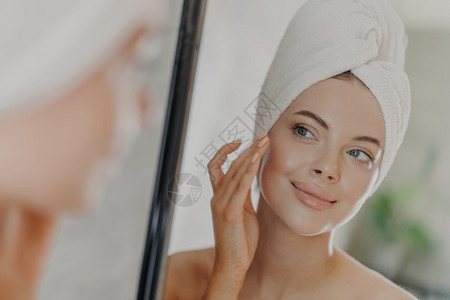 美丽的女人轻地抚摸面部看着镜子中的反光享受柔软的皮肤站在头上用毛巾裹着化妆和修甲很少美容治疗的概念图片