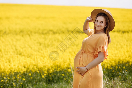 黄色种子田里的年轻孕妇。 一位年轻孕妇身着帽子和黄色太阳服的肖像。 选择性关注美丽的年轻孕妇身着黄色种子田中的年轻孕妇。图片