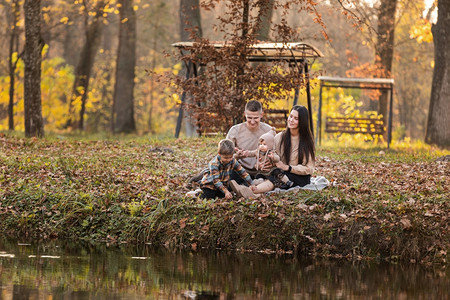 年轻爸妈和小儿子在阳光明媚的日子里在秋天公园池塘附近的毯子上玩乐幸福的家庭概念mothersfatherndsbayaydmot图片