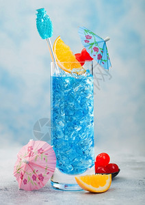 蓝色环礁湖夏季鸡尾酒在高球玻璃甜鸡尾酒樱桃和橙色切片雨伞在蓝桌背景图片