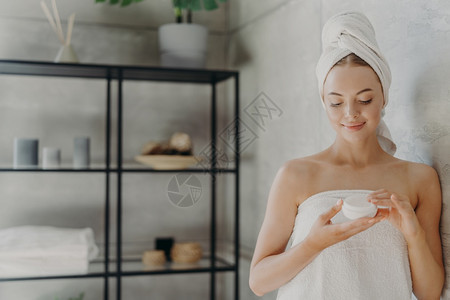 有健康皮肤的快乐女横向施用体奶油使化妆品来照顾自己在洗浴后室里摆姿势接受卫生程序图片