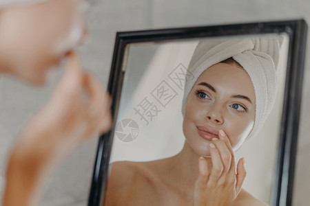 健康的妇女只化妆很少照顾皮肤和嘴唇照镜子看自己肩膀赤裸头上穿浴巾皮肤健康无瑕美貌卫生概念图片