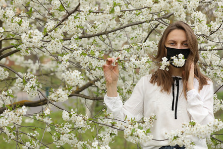 一个女孩对鲜花过敏戴着医疗面具在春日用手摸花园开的樱桃树枝一个女孩在春日用手摸花园开的樱桃树枝一个女孩在樱花开时用医疗面具对花过图片