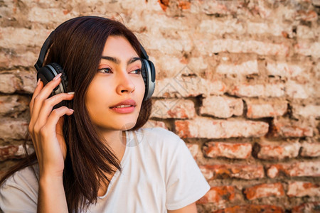 年轻拉丁女人的肖像放松听音乐用耳机对着砖墙城市概念图片