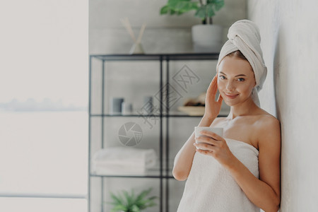 喜悦的有魅力女子自然美貌发光滑健康皮肤摸脸享受SPA程序在头上和身体周围穿白浴巾喝茶在舒适的浴室里装饰图片
