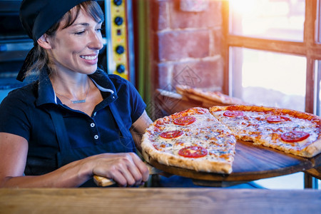穿着黑色制服的笑脸女厨师拿着木皮和披萨在比餐厅图片