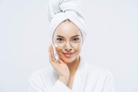 年轻自信的causin女人使用面霜享受新的反皱纹化妆品产防止皮肤衰老的迹象穿最小化妆品穿着浴袍隔离在白墙上图片