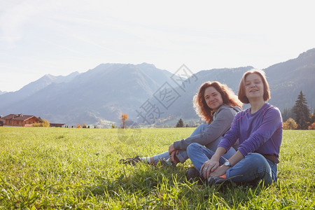 两个微笑的女孩坐在绿色草地上和山的背景图片