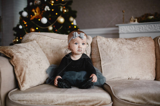 新年201和圣诞节有装饰品的圣诞树一个可爱的小女孩坐在沙发上一个穿着喜庆礼服的小孩坐在头上一个可爱的小女孩坐在圣诞树背景附近的圣图片