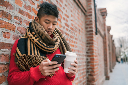 年轻亚洲人使用手机时举着咖啡杯站在街上户外图片