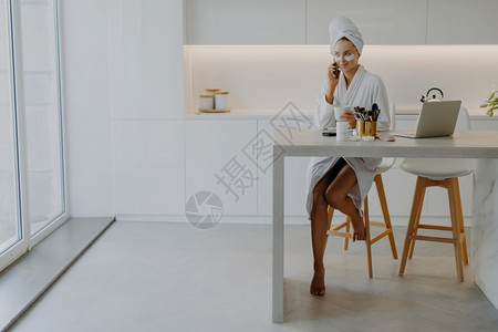 穿着浴袍的喜悦年轻女子在宽敞的厨房里赤脚坐在椅子上桌前手拿着笔记本电脑化妆品通过现代蜂窝在眼皮下交谈时使用美容修饰图片