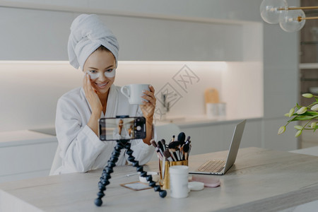 女美容博客拍摄化妆品花瓶观看智能电话谈的镜头追随者们在眼皮下使用可乐补丁喝茶时穿白色软浴袍图片