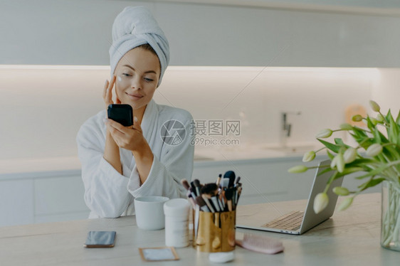 美貌佳人用面霜在脸上涂的照片镜子时看着自己关心外衣穿浴巾和袍坐在桌边对着厨房内图片