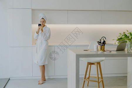 身穿白色浴袍的年轻美女 整照着脸霜镜子 顾厨房家具旁的皮肤图片