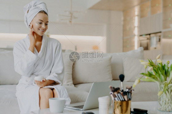 在笔记本电脑屏幕上提供穿浴袍的皮肤护理建议毛巾坐在舒适的沙发上图片