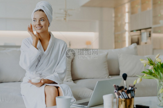 满意的女人使用面霜享受部治疗笑不看面轻使用沙发上的化妆品在开着的笔记本电脑前图片