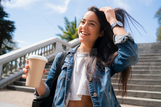 充满自信的年轻女子在户外散步时微笑并举杯咖啡的肖像城市概念图片