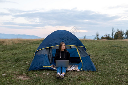 旅游妇女使用笔记本电脑在山地景观背下在帐篷附近打自由职业者字在山地景观背下旅行时打字山区有笔记本电脑的女孩旅游妇使用笔记本电脑在图片