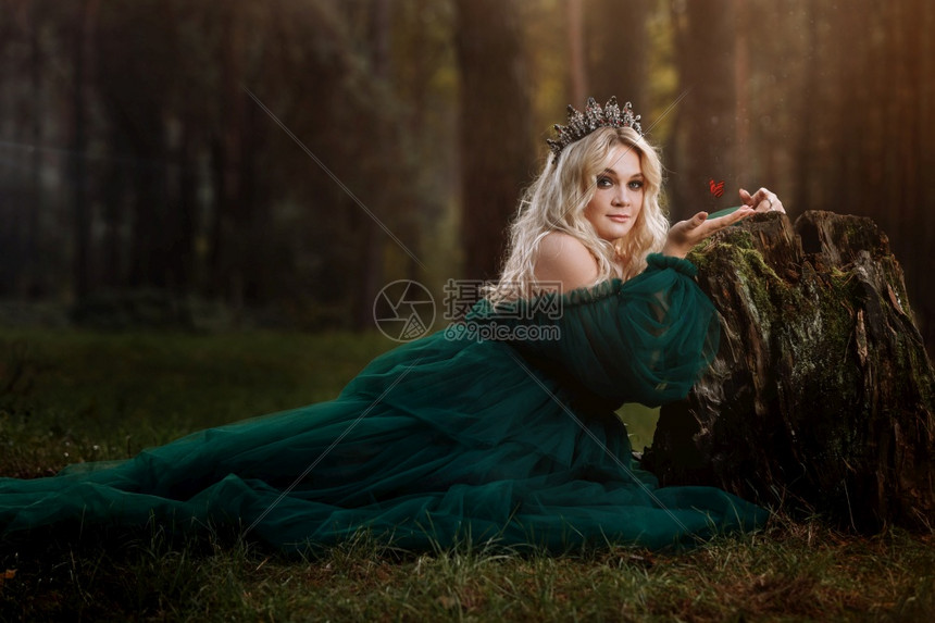 一位穿着长绿裙子的金发美女在森林中头部戴花纹坐在旧树桩旁边的女孩手拿着蝴蝶太阳光照幻想童话故事一位穿着长绿色裙子的金发美女头戴花图片