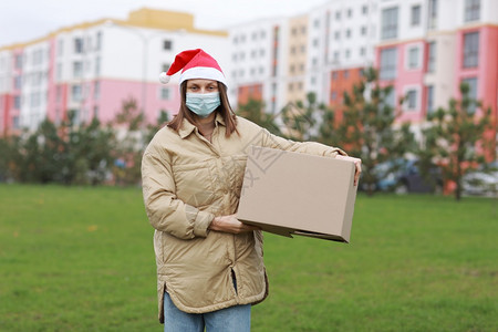 红桑塔克拉乌斯帽和医疗保护面罩的送货妇女持有一个大箱子户外新的一年和圣诞节包裹在箱内隔离时间交付线商店服务冠状红桑塔克拉乌斯帽和图片