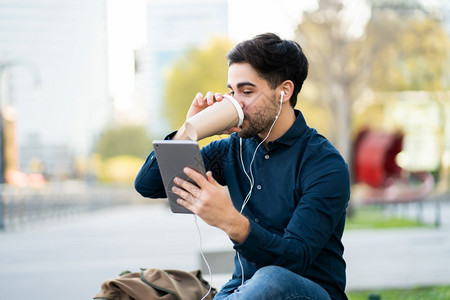 在数字平板电脑上播放视频话坐在户外的长椅上喝咖啡年轻人肖像城市概念图片