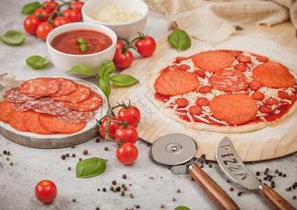 披萨和披萨刀准备烤番茄披萨配有沙拉米辣焦索配有车轮切割机新鲜西红柿和浅桌上的烤肉配有奶酪和番茄糊的碗盘背景