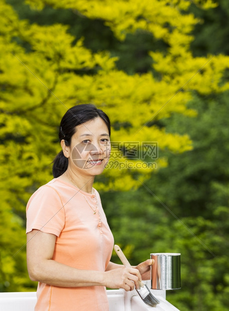 一位成年妇女在准备绿色自然背景的白木甲板上打栏时持有油漆罐和刷的垂直照片图片