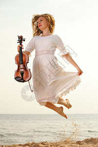 疯狂的金发女音乐情人在沙滩上骑小提琴跳跃时快乐的金发女音爱好者的概念图片