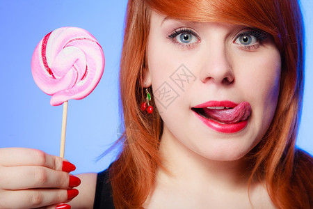 红头发女人感的年轻女人拿着糖果舔她的嘴唇红头发女孩在紫罗兰上吃甜的棒糖背景