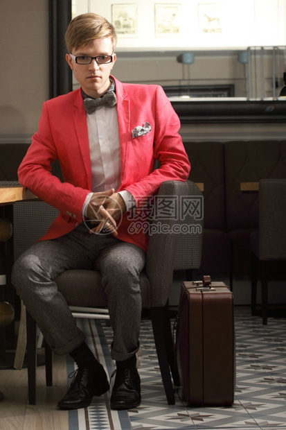 年轻帅的时装模特身着眼镜的年轻时装帅男时装模特身着明亮红色夹克领结带和手提箱在室内等候图片