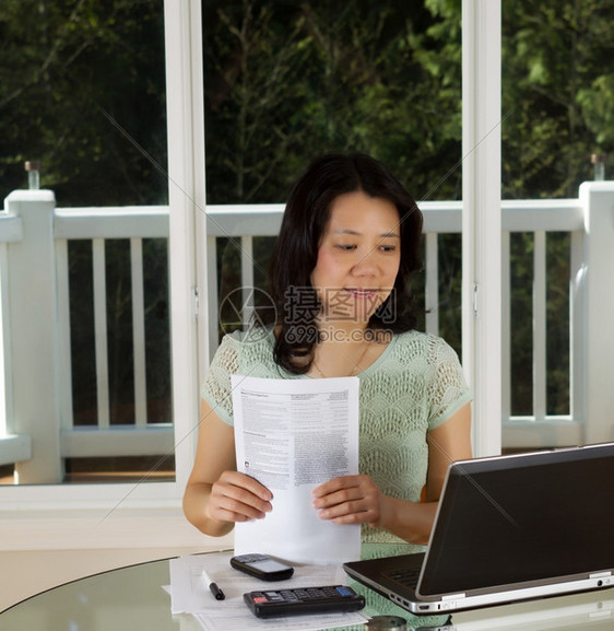 成年妇女看计算机屏幕同时持有税表在家用笔记本电脑计算器和纸在桌顶大背景窗口上工作的照片图片