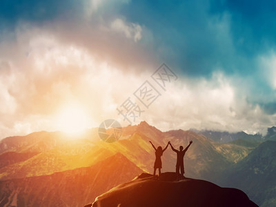 一对幸福的情侣站在山峰的上双手举起在日落时抬起令人叹为观止的美景图片
