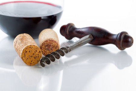 紧近古董corkscrew前视聚焦于corkscrew的前部和远左侧cork放在玻璃桌上杯里有红葡萄酒背景与反射图片