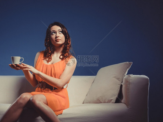 长卷发女孩时的橙色裙子拿着咖啡茶杯热饮坐在沙发上望向室内图片