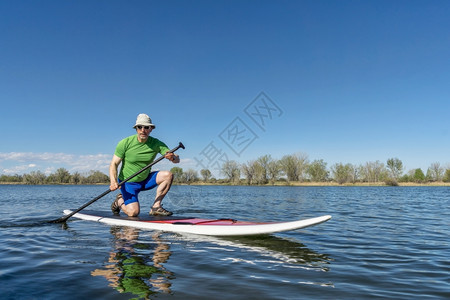 时尚的老人在平静的湖中间跪在皮划桨上划水图片