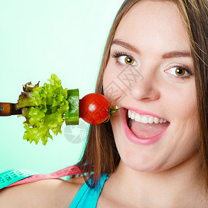 健美的身女拿着新鲜混合蔬菜的叉子和蓝色背景的测量胶带图片