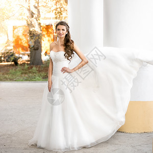 穿着长白色裙子的美丽黑发女新娘假扮在公园风天背景图片