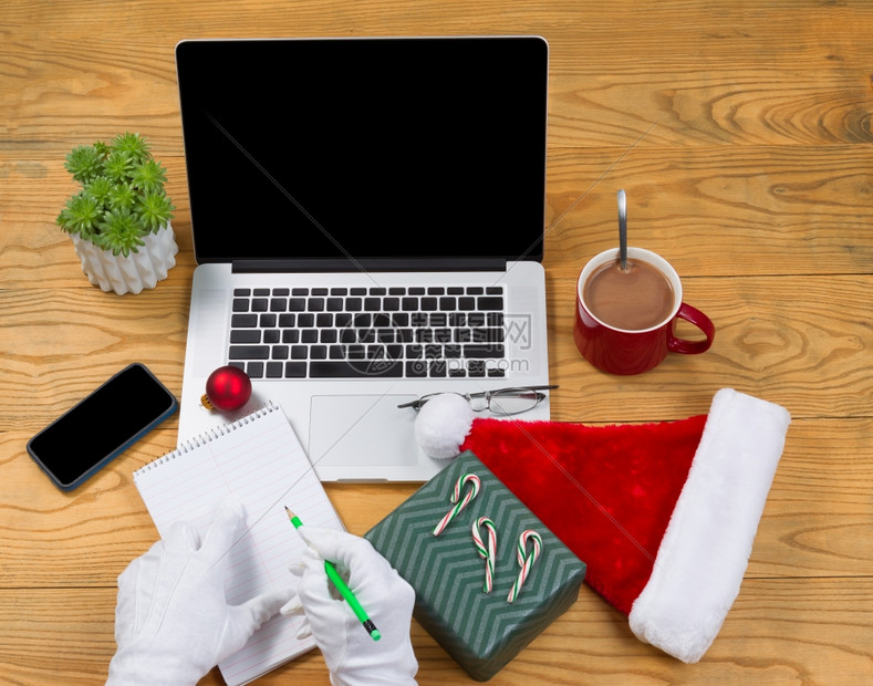 圣诞老人用热巧克力电脑现货帽子笔记本铅手机阅读眼镜和桌面上的植物来写礼清单圣诞老人办公室的概念图片