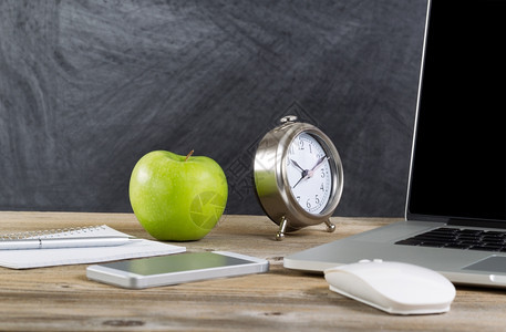 在黑板前装有笔记本电脑鼠标时钟手机笔记本和绿苹果的学校桌面图片