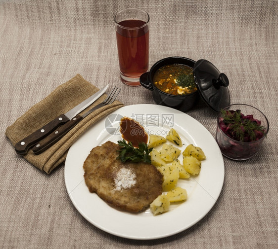 PetiteWienerschnitzel配有煮土豆和番茄酱身着白色瓷板用叉子和刀在木制背景上甜菜沙拉和番茄红辣椒汤橄榄油酱迷迭图片