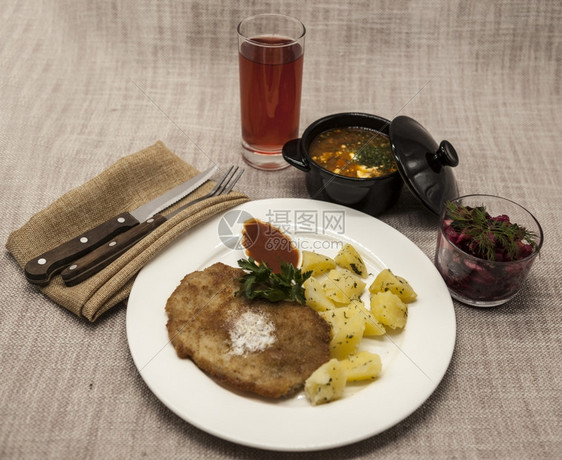 PetiteWienerschnitzel配有煮土豆和番茄酱身着白色瓷板用叉子和刀在木制背景上甜菜沙拉和番茄红辣椒汤橄榄油酱迷迭图片