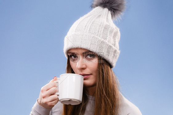 穿着冬羊毛帽的有吸引力长发女郎拿着杯子喝茶热饮给自己工作室暖身图片