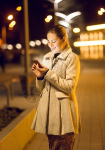 美丽的年轻女孩晚上在街使用智能手机图片