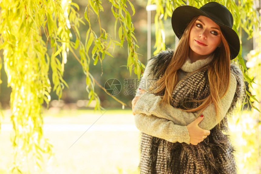 穿着时髦的毛衣和露天热帽的年轻美女站在柳树叶旁的公园里图片