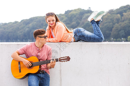 吉他主义者和的穆斯爱浪漫音乐天才激情约会概念吉他主义者和的缪斯年轻男子与躺在墙上有风景背的女孩一起弹吉他背景图片