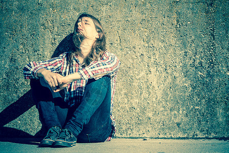 长的头发独自坐在墙壁上悲伤的长发男人的头发独自在户外的墙上悲伤长发男人失业抑郁或悲伤的概念图片