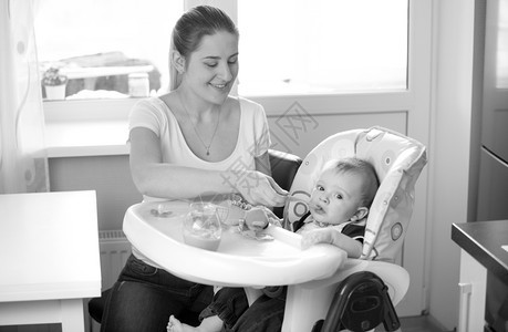 年轻笑母亲在厨房用勺子喂婴儿的黑白画像图片