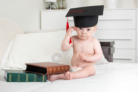 可爱的10个月大男孩坐在床上带着书和并脱下毕业上限图片